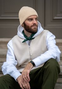 Γκρίζα φλις γιλέκο πάνω από λευκή φόρμα, πράσινο μαντίλι, χακί παντελόνι και ανοιχτό καφέ ριχτό σκουφάκι; καθισμένος στα σκαλιά, χαλαρή στάση.