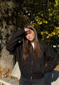 Sweat à capuche noir avec fermeture éclair, coupe décontractée, poignets côtelés et poches avant. Porté sur une chemise blanche, avec des cheveux longs et raides, sous la lumière naturelle du soleil.