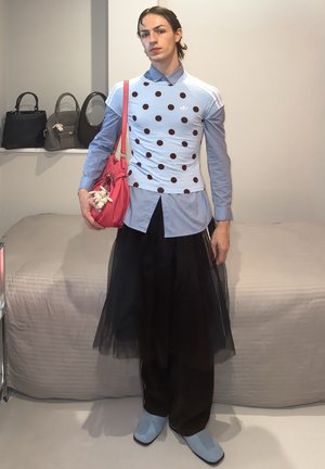 Persona que lleva una camisa azul clara con lunares encima de una camisa azul con cuello, falda de tul negro sobre pantalones, zapatos azul claro, sosteniendo un bolso rosa con llaveros.