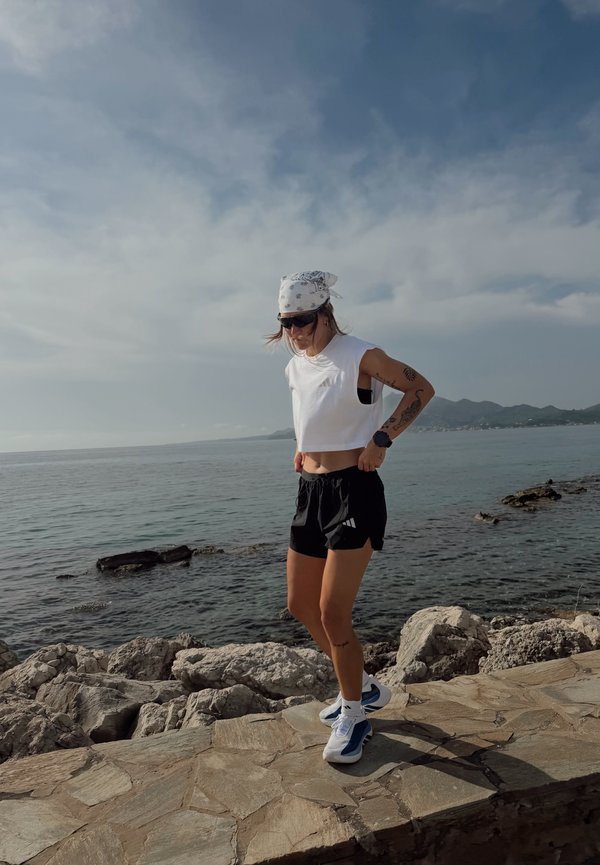 Vrouw in sportkleding staat op een stenen pad aan zee, draagt een zonnebril, bandana en hardloopschoenen op een gedeeltelijk bewolkte dag.