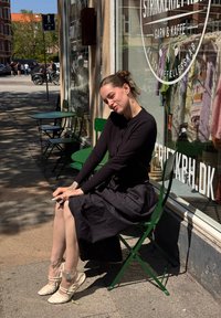 Fekete ruhás nő ül egy zöld fém széken egy fonal- és kávézó előtt, mögötte kötött pulcsik vannak kiállítva az ablakban.