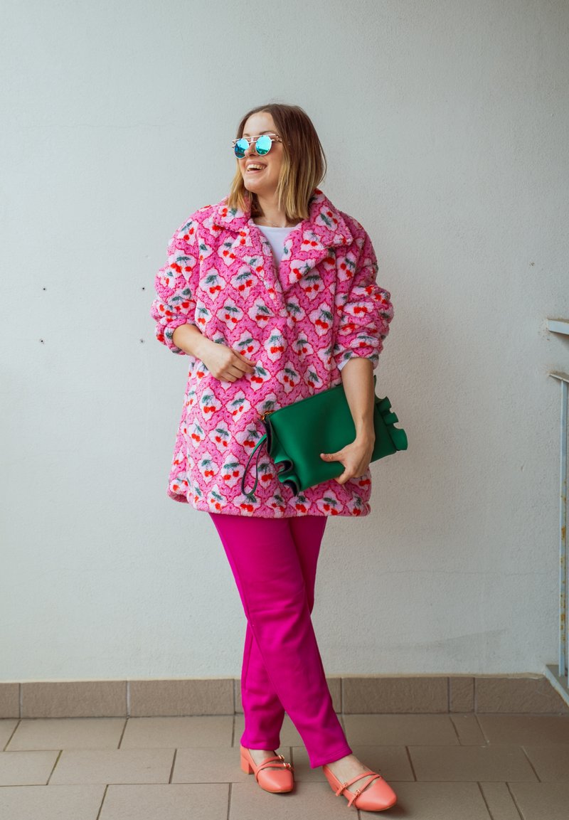 Femme portant des lunettes de soleil réfléchissantes, un manteau à fleurs roses, un pantalon rose vif, des chaussures corail, tenant une pochette verte, debout contre un mur uni.