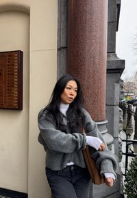 Jeune femme aux longs cheveux noirs portant des pulls gris superposés et un jean noir, appuyée contre une colonne en pierre rouge en plein air.
