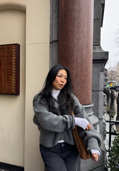 Joven con cabello largo y negro, vistiendo suéteres grises en capas y jeans negros, recostada contra una columna de piedra roja al aire libre.