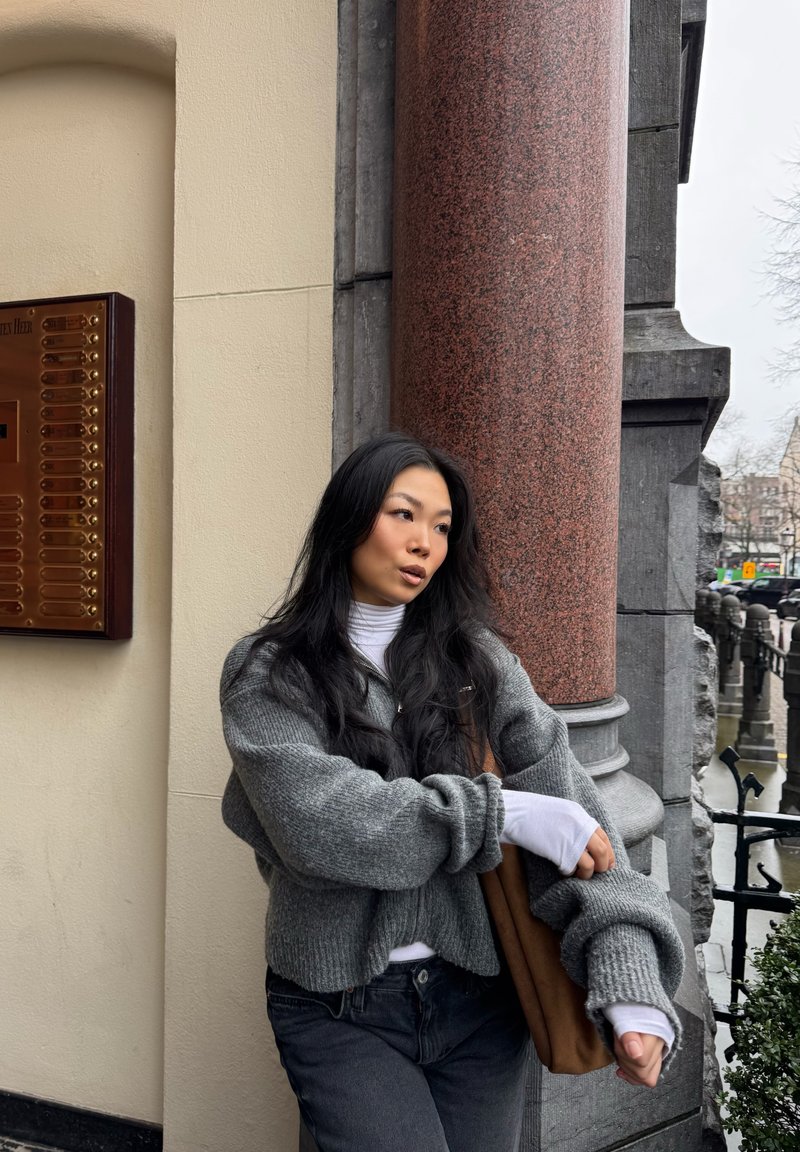Giovane donna con capelli neri lunghi che indossa maglioni grigi a strati e jeans neri, appoggiata a una colonna di pietra rossa all'aperto.