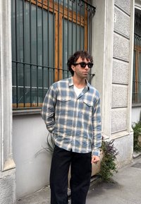 A blue and white plaid button-up shirt featuring two chest pockets, teamed with black trousers. The wall behind has a grey texture with wooden window frames.