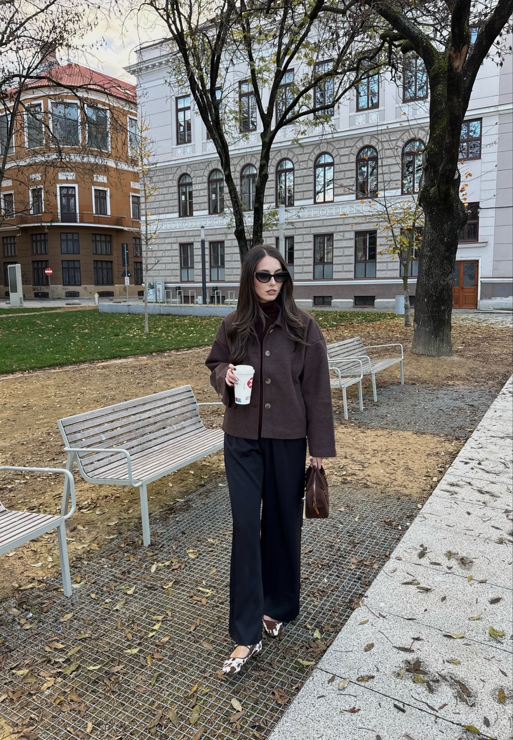 Mörkbrun jacka med knappar, svarta vida byxor och ballerinaskor med kohudsmönster. Håller i en kaffekopp och promenerar i en park med bänkar och byggnader.