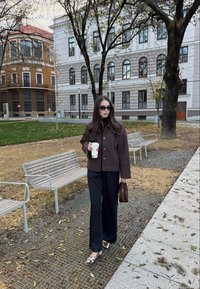 Mörkbrun jacka med knappar, svarta vida byxor och ballerinaskor med kohudsmönster. Håller i en kaffekopp och promenerar i en park med bänkar och byggnader.