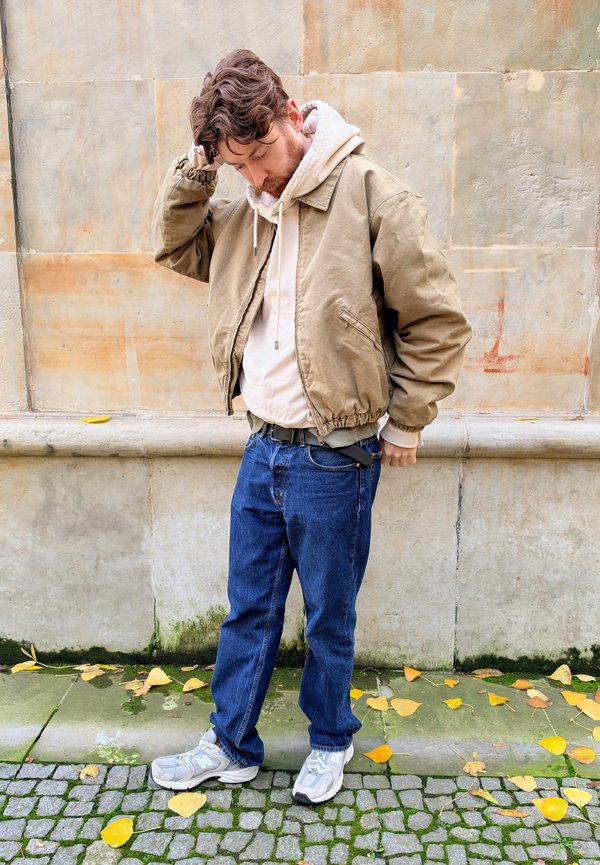 Un homme portant une veste beige, un sweat à capuche blanc, un jean bleu et des baskets grises se recoiffe en se tenant près d’un mur en pierre.