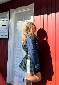 Μοντέλο φοράει ένα τζάκετ σε ναυτικό μπλε με καρό σχέδιο και ρίγες, σε συνδυασμό με μια καρό φούστα, μπροστά από έναν κόκκινο ξύλινο τοίχο κα�ι μια λευκή πόρτα.