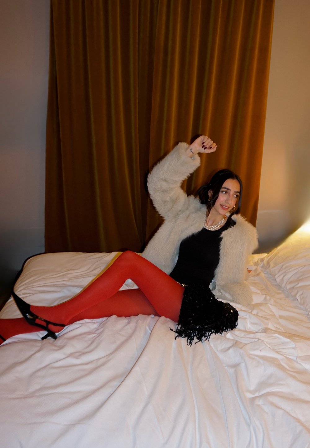Casaco felpudo branco, blusa preta ajustada, saia mini preta com lantejoulas, collants vermelhos e saltos pretos sobre uma cama com roupa de cama branca amassada e cortinas marrons.