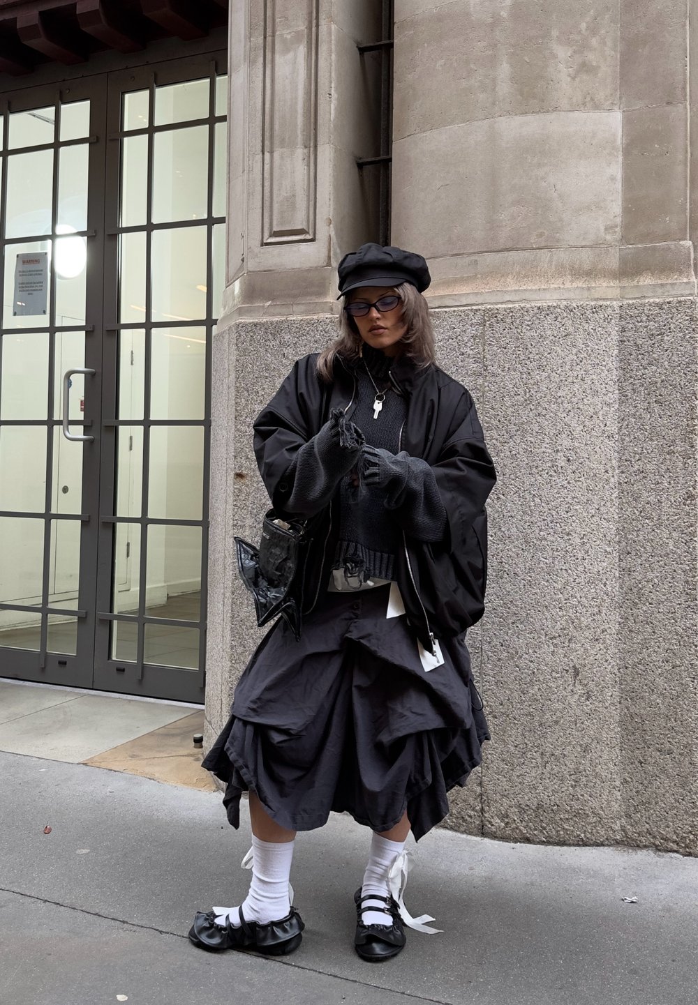 Čierna nadmerná bunda, vrstvená čierna sukňa s volánmi, pletený sveter, čierna taška a topánky s bielymi stuhami. Doplnky zahŕňajú okuliare a čiapku.