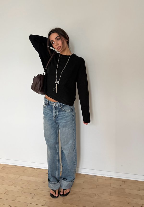 Mujer de pie contra una pared lisa, vestida con un suéter negro, vaqueros azules holgados, chanclas, un collar largo y llevando un bolso marrón colgado del hombro.