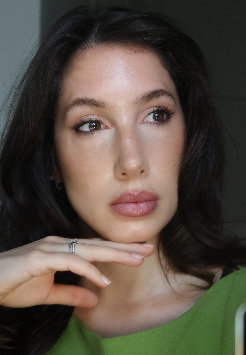 Femme aux cheveux bruns et top vert, reposant son menton sur sa main, portant une bague en argent, regardant pensivement sur le côté.