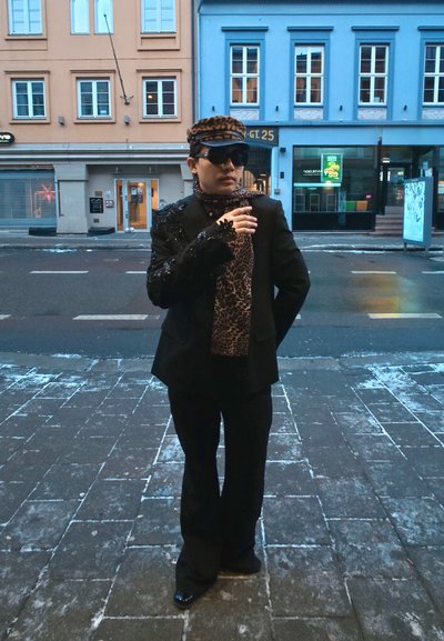 Traje negro con hombrera adornada, bufanda de estampado de leopardo, gafas de sol y gorra a juego. Acera y edificios al fondo.