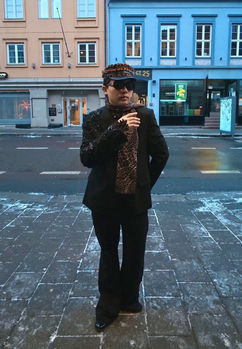 Costume noir avec épaule ornée, foulard imprimé léopard, lunettes de soleil et casquette assortie. Trottoir et bâtiments en arrière-plan.