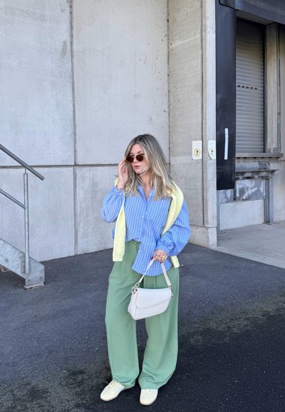 Mujer con camisa azul a rayas, pantalones anchos verdes, jersey amarillo sobre los hombros, gafas de sol, sosteniendo bolso blanco, de pie junto a una pared de hormigón.