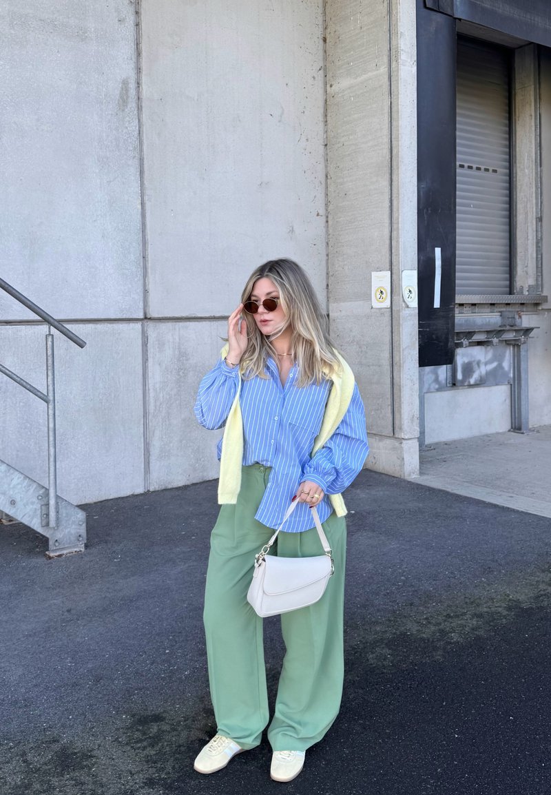 Moteris vilki mėlyną dryžuotą marškinius, žalias plačias kelnes, geltoną megztinį per pečius, dėvi akinius nuo saulės, laiko baltą rankinę, stovi prie betono sienos.