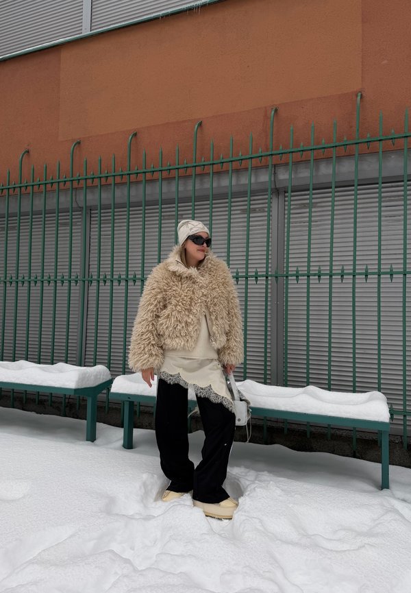 Donna con giacca beige pelosa, pantaloni neri, berretto crema e occhiali da sole in piedi sulla neve vicino a una recinzione metallica verde e panchine con neve.