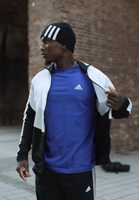 Adidas juoda ir balta striukė užmesta ant mėlyno marškinėlio, abu su logotipu. Juoda kepurė su baltomis juostelėmis. Miestinis plytų fonas.