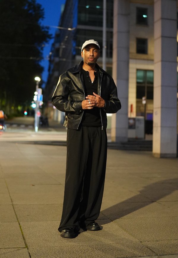 Hombre con chaqueta de cuero negra, pantalones negros y gorra blanca, de pie en una calle de la ciudad por la noche con edificios y luces de fondo.