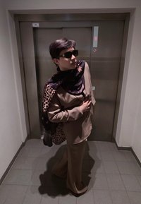 Personne portant un costume marron et une écharpe à motifs, posant la main sur la poitrine devant des portes d'ascenseur fermées dans un couloir carrelé.