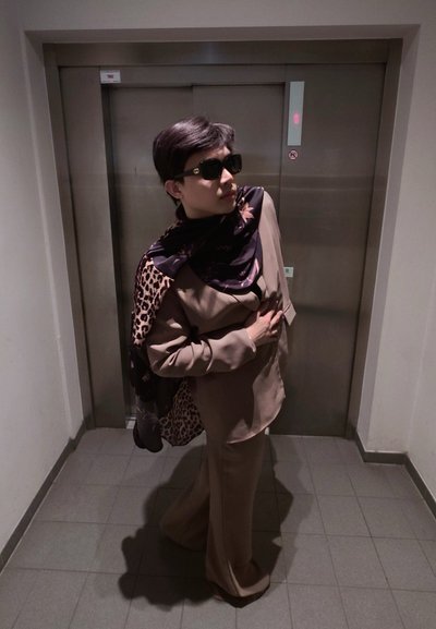 Persona con traje marrón y bufanda estampada, posando con la mano en el pecho frente a unas puertas de ascensor cerradas en un pasillo con azulejos.