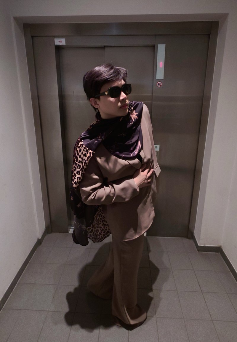 Personne portant un costume marron et une écharpe à motifs, posant la main sur la poitrine devant des portes d'ascenseur fermées dans un couloir carrelé.