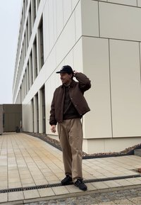 Veste pilote marron portée sur un pull texturé, pantalon beige et chaussures noires. Debout à l'extérieur près d'un bâtiment moderne avec un motif en grille.