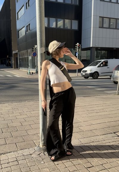 Persona que lleva una camiseta corta blanca y pantalones amplios negros, sosteniendo un teléfono. Una gorra beige y un cinturón con hebilla completan el look.