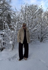 Manteau en fausse fourrure beige clair, coupe oversize ; associé à un pantalon large foncé et des chaussures marron clair ; cadre hivernal avec des arbres enneigés.