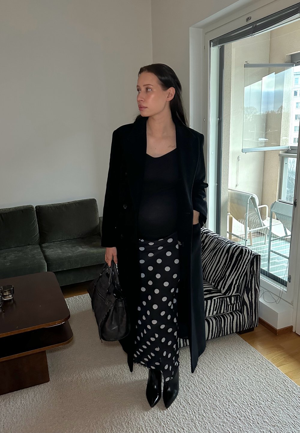 Fekete gyapjú kabát fekete, testhezálló felsővel, pöttyös szoknyával és hegyes orrú fekete csizmával párosítva. Egy texturált fekete kézitáskát tart a kezében.