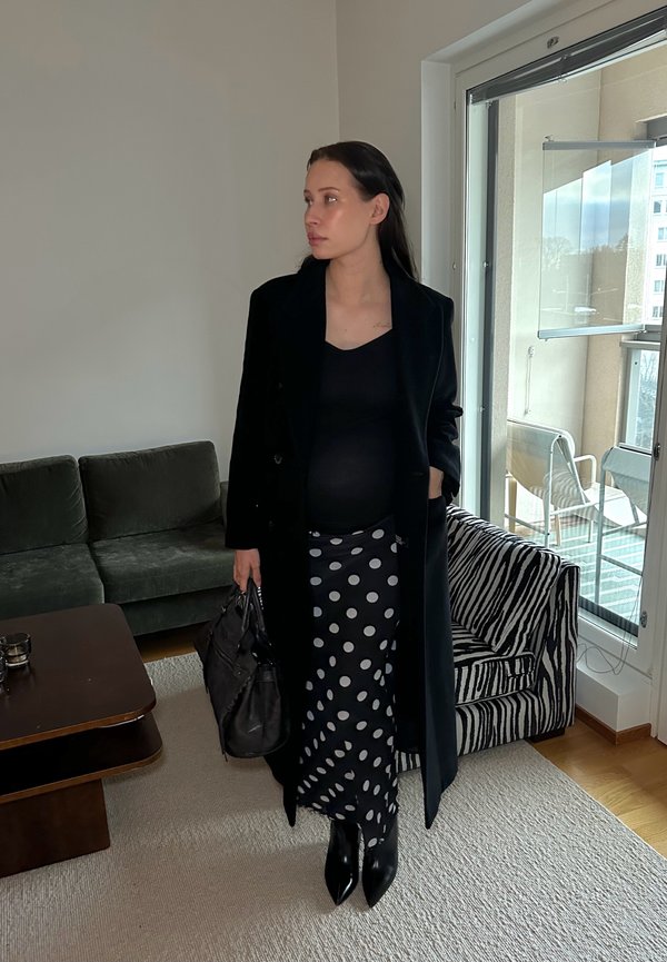 Fekete gyapjú kabát fekete, testhezálló felsővel, pöttyös szoknyával és hegyes orrú fekete csizmával párosítva. Egy texturált fekete kézitáskát tart a kezében.