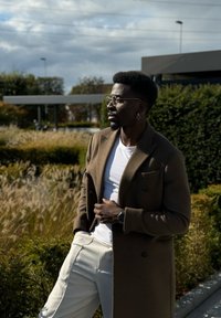 Καφέ μάλλινο παλτό με μεγάλα καπέλα, φορεμένο πάνω από ένα λευκό μπλουζάκι. Ανοιχτόχρωμο παντελόνι και στρογγυλά γυαλιά, σε έναν χλοώδη εξωτερικό χώρο.