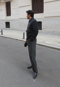 Giacca di lana scura con colletto strutturato, pantaloni grigi su misura e scarpe nere lucide, completati da guanti neri. Ambientazione urbana in strada.