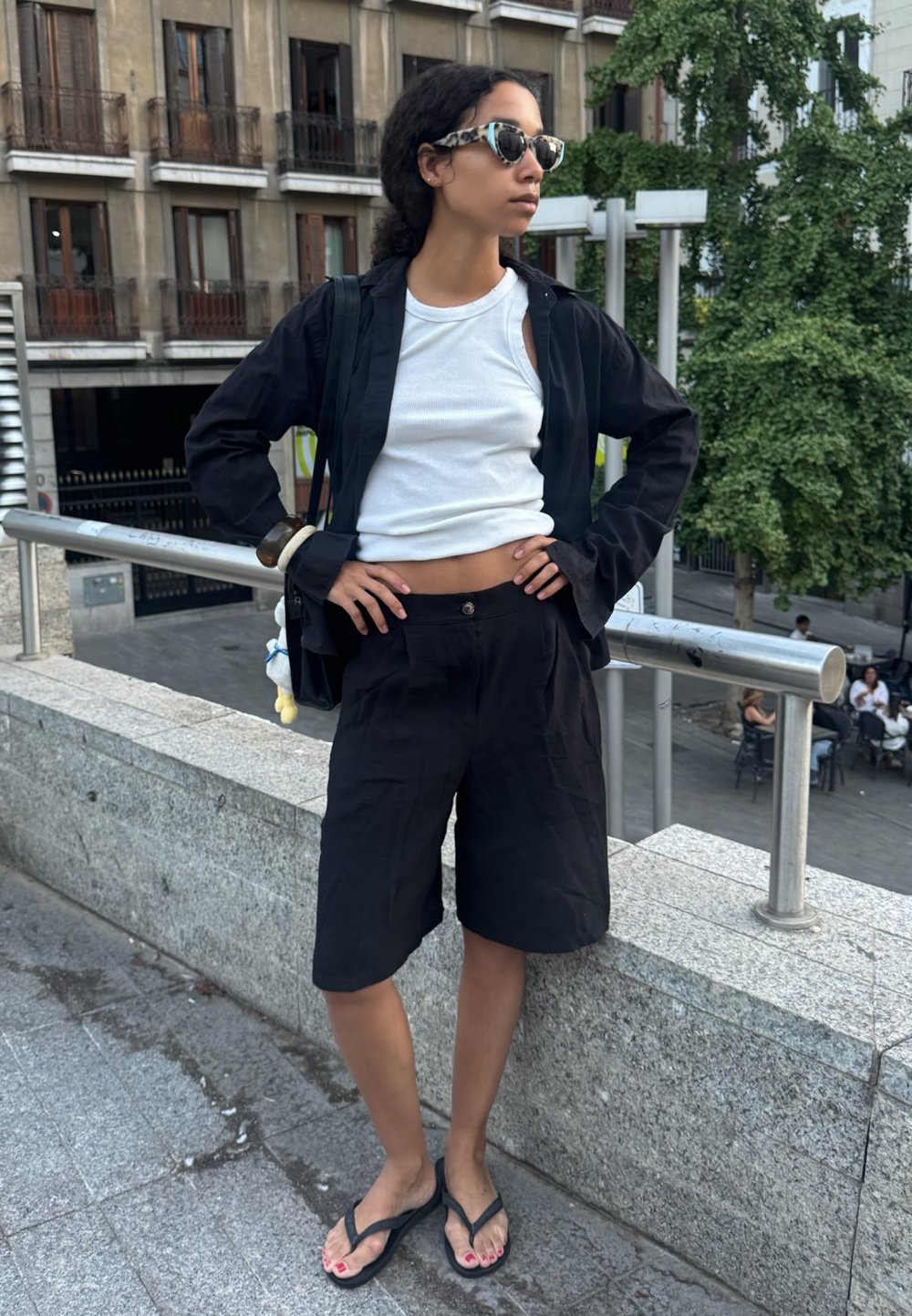 Vrouw in zonnebril, gekleed in een witte tanktop, een zwarte open blouse, zwarte shorts en slippers, staat met de handen op de heupen op een stedelijke stenen rand.