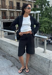 Vrouw in zonnebril, gekleed in een witte tanktop, een zwarte open blouse, zwarte shorts en slippers, staat met de handen op de heupen op een stedelijke stenen rand.
