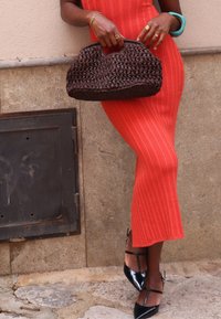 A ribbed, form-fitting orange dress reaches below the knee. A woven dark brown handbag is held in one hand, paired with shiny black heels.
