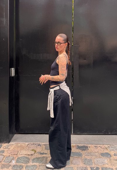 Camiseta negra sin mangas, pantalones anchos negros con una corbata gris en la parte de atrás, y gafas. Brazos tatuados. De pie contra una pared negra. Suelo de adoquines.