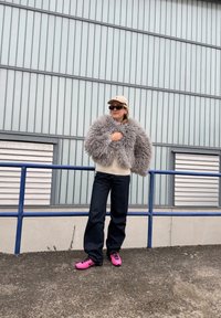 Personne debout à l'extérieur portant une casquette beige, des lunettes de soleil foncées, une veste grise duveteuse, un pull crème, un jean foncé et des baskets roses vives près d'une rambarde en métal.