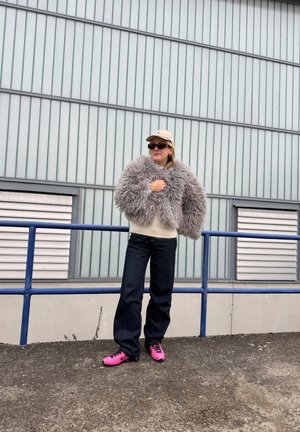 Persona de pie al aire libre con gorra beige, gafas de sol oscuras, chaqueta gris esponjosa, suéter crema, vaqueros oscuros y zapatillas de deporte rosa brillante junto a una barandilla metálica.