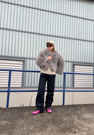 Persona de pie al aire libre con gorra beige, gafas de sol oscuras, chaqueta gris esponjosa, suéter crema, vaqueros oscuros y zapatillas de deporte rosa brillante junto a una barandilla metálica.