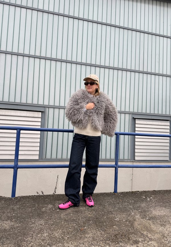 Person som står utendørs iført beige caps, mørke solbriller, grå fluffy jakke, kremhvit genser, mørke jeans og sterke rosa joggesko ved metallrekkverk.