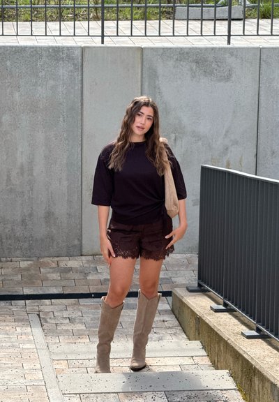 Joven mujer con el pelo largo y ondulado está de pie en unos escalones pavimentados, lleva un top negro y pantalones cortos, botas beige hasta la rodilla y un bolso beige de hombro.