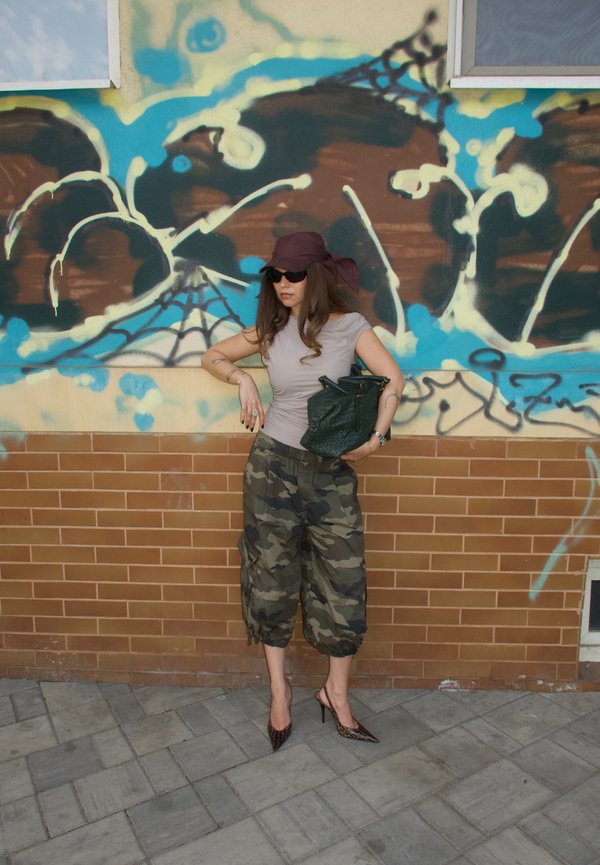 Kvinde iført solbriller, lilla hat, beige top og camouflagebukser holder en grøn taske og står mod en murstensvæg med graffiti.