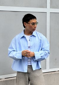 Világoskék, túlméretezett ing fehér függőleges csíkokkal, fehér alsó inggel és világosszürke nadrággal párosítva; laza gallérral és gombos megoldással.