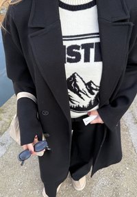 Manteau en laine noir à la coupe ajustée, pull blanc avec motif de montagnes, pantalon noir à jambes larges et chaussures beiges à enfiler.