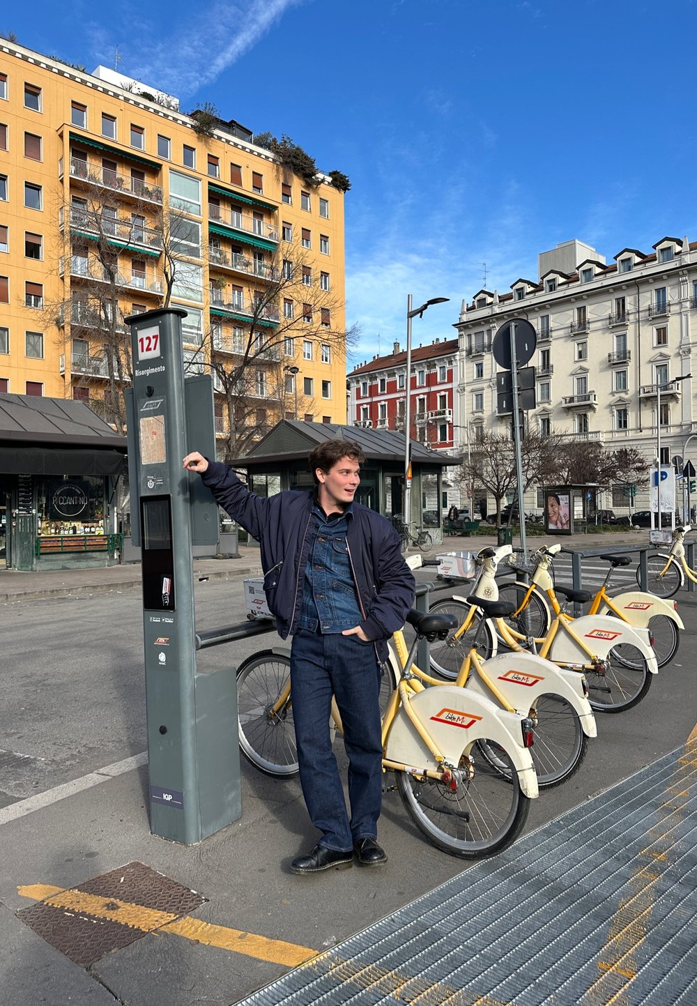 Σταθμός ενοικίασης ποδηλάτων με γκρι περίπτερο, κίτρινα ποδήλατα με άσπρους στάντ, και ένα νέο άτομο με σκούφο και τζιν.