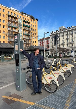 Estación de alquiler de bicicletas con kioscos grises, bicicletas amarillas con estantes blancos, y una persona joven con chaqueta oscura y vaqueros.