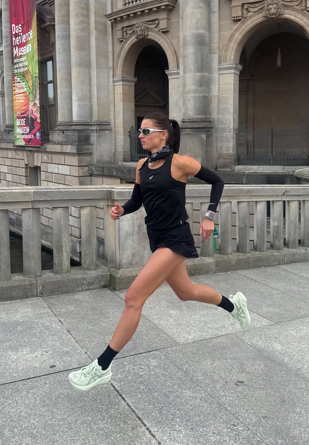 Coureur en débardeur noir et shorts, portant des manchons de bras noirs et des chaussures verdâtres, courant sur un chemin en pierre avec en toile de fond un musée.